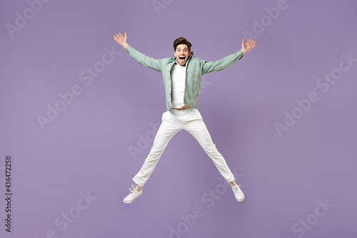 Full length fun excited overjoyed amazed cool young man in casual mint shirt white t-shirt jump high with outstretched hands look camera isolated on purple violet background. People lifestyle concept © ViDi Studio