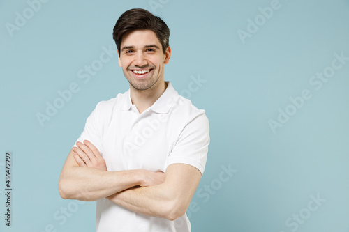 Young fun smiling happy unshaven cheerful caucasian man 20s wear white casual basic t-shirt hold hands crossed folded isolated on pastel blue color background studio portrait People lifestyle concept
