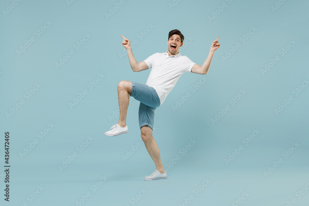 Full length young fun happy overjoyed man 20s wear white basic t-shirt do winner gesture point finger with raised up leg isolated on pastel blue background studio portrait. People lifestyle concept