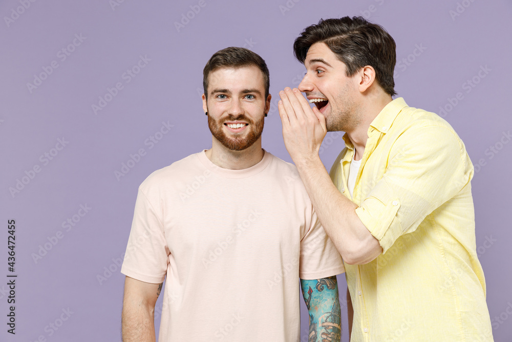 Naklejka premium Two smiling young men friends together wear casual t-shirt tattoo translate fun whispering gossip and tells secret behind his hand sharing news isolated on purple background. People lifestyle concept.