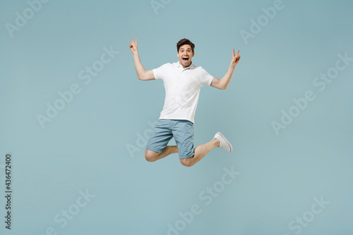 Full length smiling young man 20s wear white casual basic t-shirt showing victory v-sign gesture jump high looking camera isolated on pastel blue background studio portrait. People lifestyle concept.