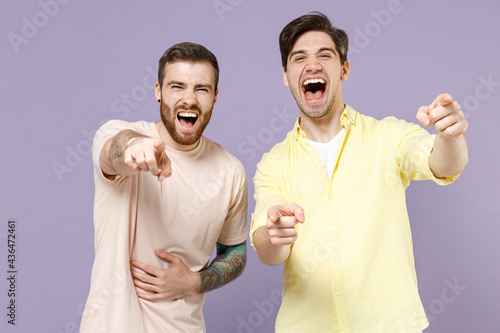 Two smiling amazed happy young men 20s friends together wearing casual t-shirt point index fingers camera on you laughing mocking joking isolated on purple background studio People lifestyle concept
