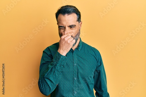 Middle age man with beard and grey hair wearing business clothes smelling something stinky and disgusting  intolerable smell  holding breath with fingers on nose. bad smell