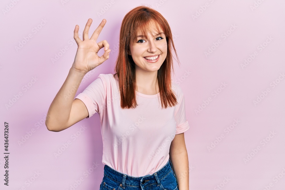 Redhead young woman wearing casual pink t shirt smiling positive doing ok sign with hand and fingers. successful expression. Redhead young woman wearing casual pink t shirt smiling positive doing ok sign with hand and fingers. successful expression.
