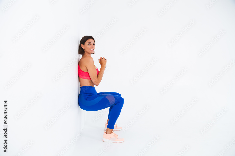 Fit tanned sporty woman with abs, fitness curves, wearing top and blue leggings on white background does chair exercise
