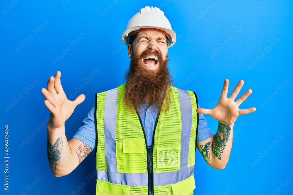 Redhead man with long beard wearing safety helmet and reflective jacket crazy and mad shouting and yelling with aggressive expression and arms raised. frustration concept.