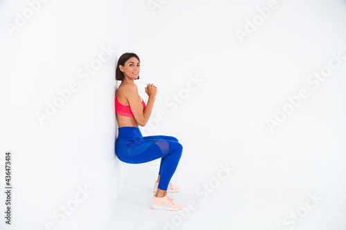 Fit tanned sporty woman with abs, fitness curves, wearing top and blue leggings on white background does chair exercise