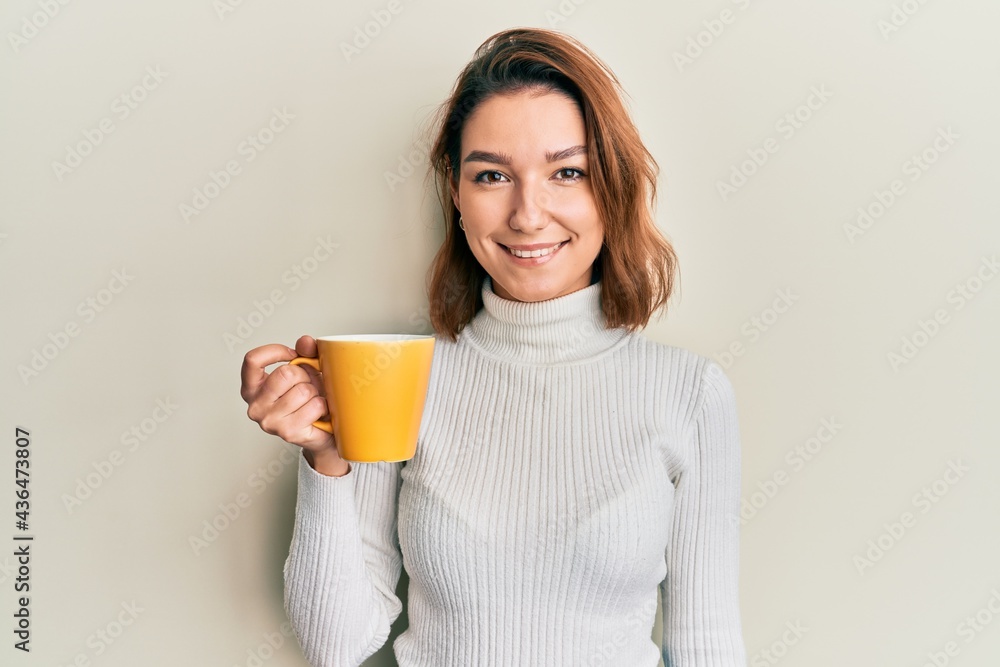 Young caucasian woman holding coffee looking positive and happy standing and smiling with a confident smile showing teeth Young caucasian woman holding coffee looking positive and happy standing and smiling with a confident smile showing teeth