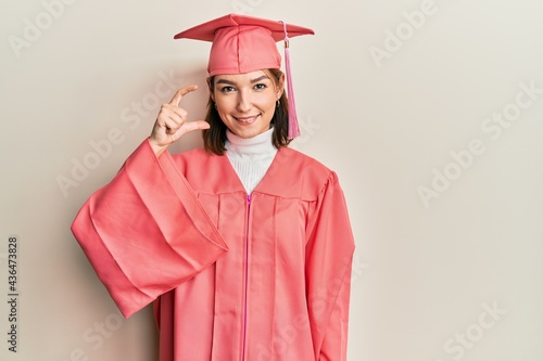 Young caucasian woman wearing graduation cap and ceremony robe smiling and confident gesturing with hand doing small size sign with fingers looking and the camera. measure concept.