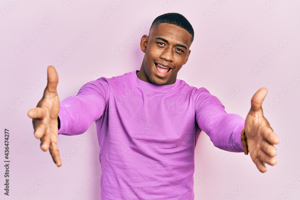 Young black man wearing casual pink sweater looking at the camera smiling with open arms for hug. cheerful expression embracing happiness. Young black man wearing casual pink sweater looking at the camera smiling with open arms for hug. cheerful expression embracing happiness.
