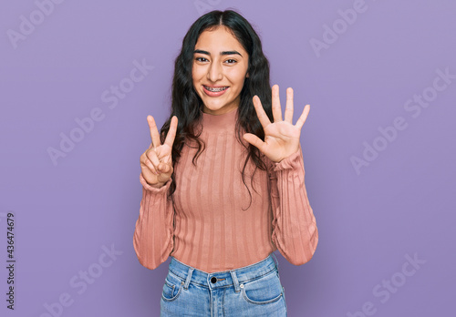 Hispanic teenager girl with dental braces wearing casual clothes showing and pointing up with fingers number seven while smiling confident and happy.