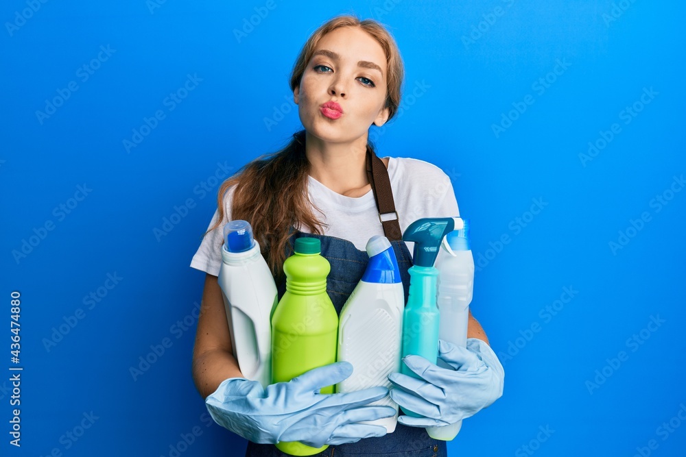 Beautiful blonde caucasian woman wearing cleaner apron holding cleaning products looking at the camera blowing a kiss being lovely and sexy. love expression. Beautiful blonde caucasian woman wearing cleaner apron holding cleaning products looking at the camera blowing a kiss being lovely and sexy. love expression.