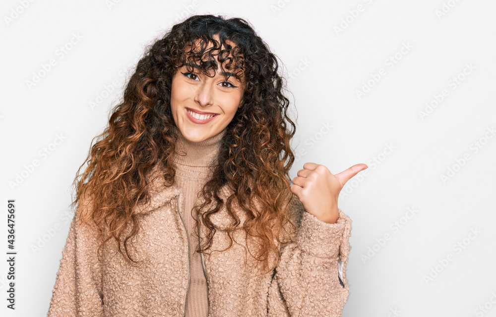 Young hispanic girl wearing winter clothes smiling with happy face looking and pointing to the side with thumb up. Young hispanic girl wearing winter clothes smiling with happy face looking and pointing to the side with thumb up.