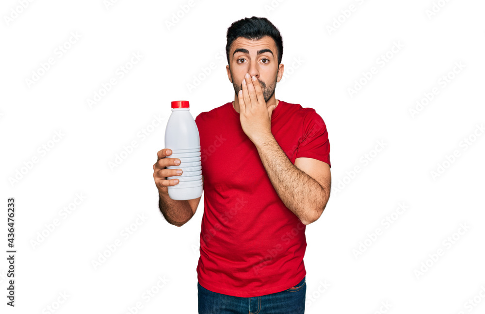 Hispanic man with beard holding liter bottle of milk covering mouth with hand, shocked and afraid for mistake. surprised expression Hispanic man with beard holding liter bottle of milk covering mouth with hand, shocked and afraid for mistake. surprised expression