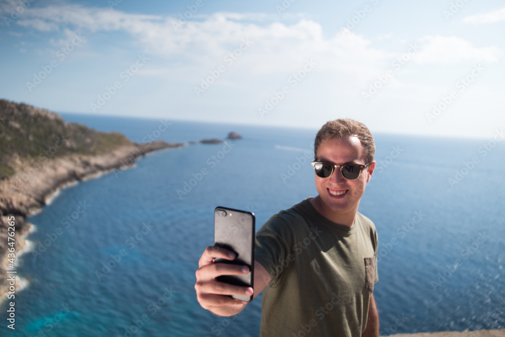 Happy young adult smiling and wearing sunglasses holding a smartphone and taking a selfie with the ocean in the background. 
