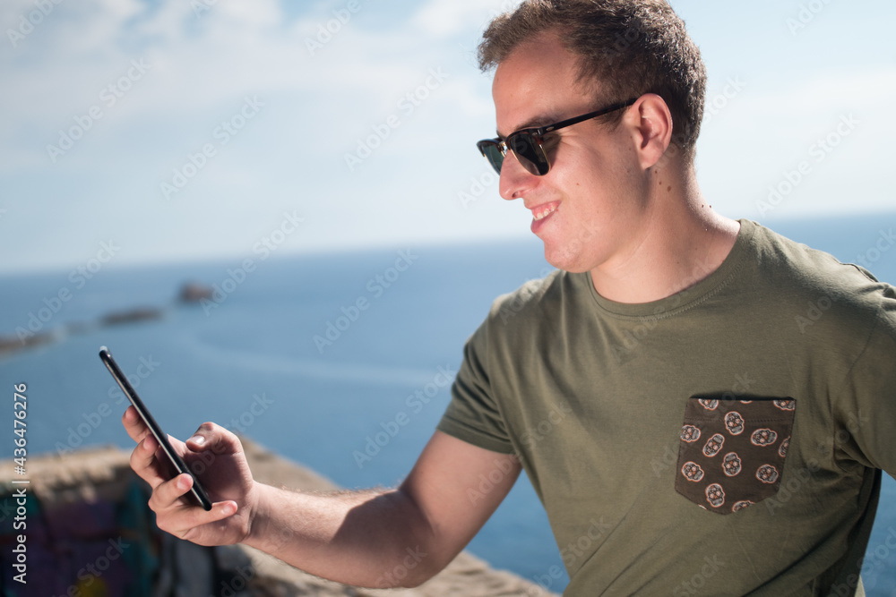 Young adult smiling and wearing sunglasses holding an smartphone with the ocean in the background. 