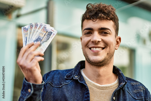 Young hispanic man smiling happy holding peruvian sol banknotes at the city