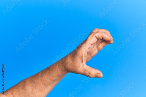 Hand of senior hispanic man over blue isolated background picking and taking invisible thing, holding object with fingers showing space