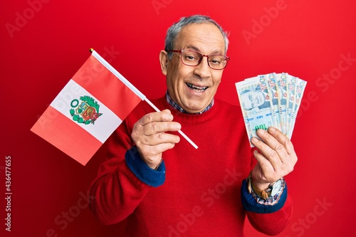 Handsome senior man with grey hair holding peru flag and peruvian sol banknotes smiling and laughing hard out loud because funny crazy joke.