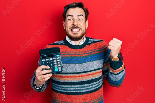 Handsome caucasian man with beard showing calculator device screaming proud, celebrating victory and success very excited with raised arm