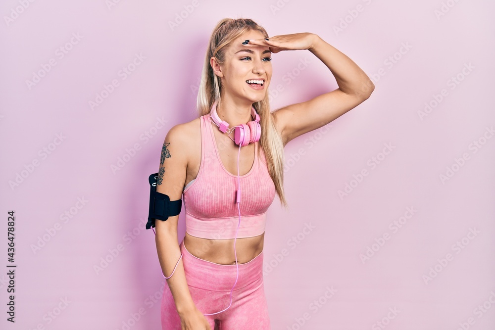 Obraz premium Young beautiful caucasian woman wearing gym clothes and using headphones very happy and smiling looking far away with hand over head. searching concept.