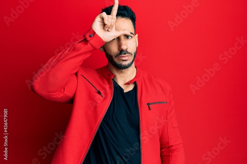 Young hispanic man wearing red leather jacket making fun of people with fingers on forehead doing loser gesture mocking and insulting. © Krakenimages.com