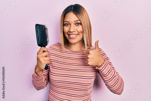 Beautiful hispanic woman holding hairbrush smiling happy and positive, thumb up doing excellent and approval sign