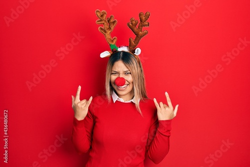 Beautiful hispanic woman wearing deer christmas hat and red nose shouting with crazy expression doing rock symbol with hands up. music star. heavy concept.