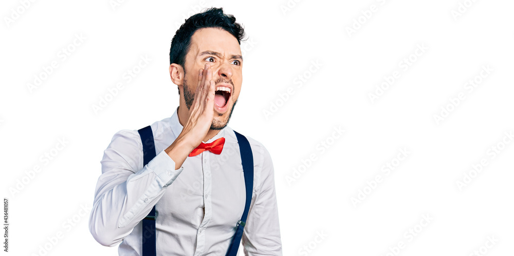 Hispanic man with beard wearing hipster look with bow tie and suspenders shouting and screaming loud to side with hand on mouth. communication concept. Hispanic man with beard wearing hipster look with bow tie and suspenders shouting and screaming loud to side with hand on mouth. communication concept.