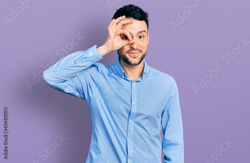 Hispanic man with beard wearing casual business shirt doing ok gesture with hand smiling, eye looking through fingers with happy face.