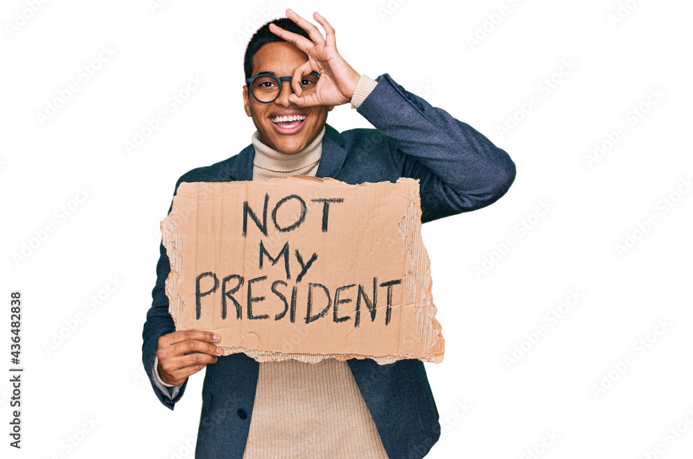 Young handsome hispanic man holding not my president protest banner smiling happy doing ok sign with hand on eye looking through fingers Young handsome hispanic man holding not my president protest banner smiling happy doing ok sign with hand on eye looking through fingers