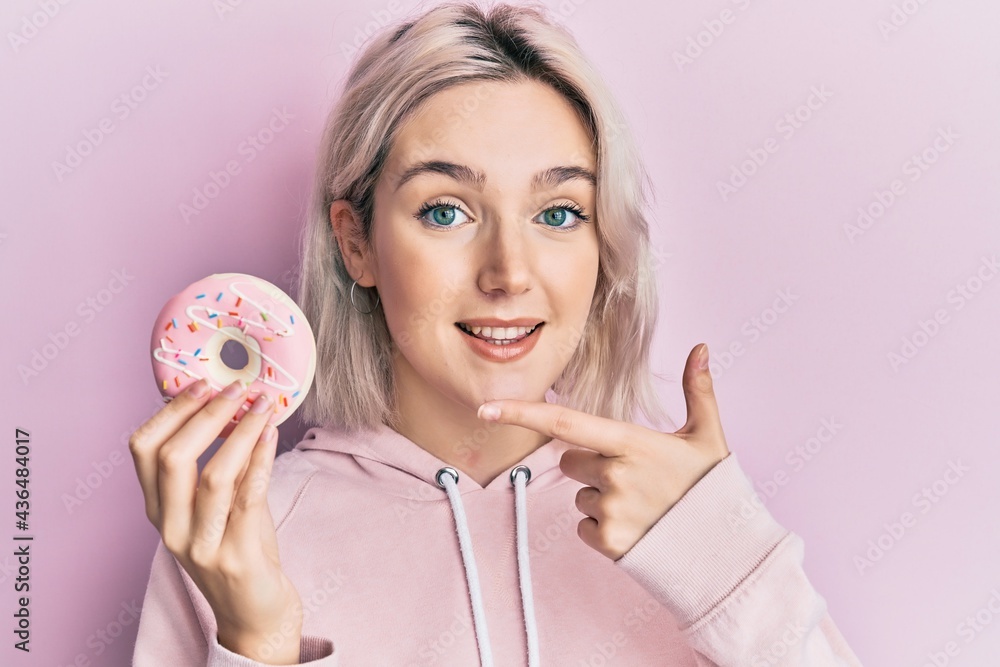 Young blonde girl holding donut smiling happy pointing with hand and finger Young blonde girl holding donut smiling happy pointing with hand and finger