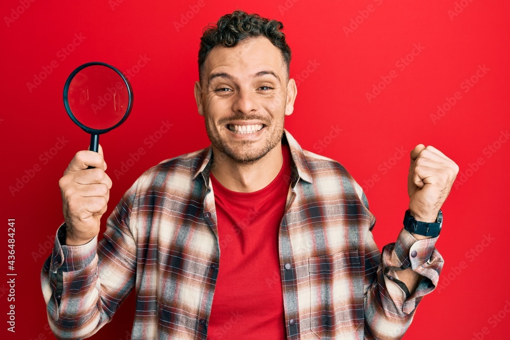Young hispanic man holding magnifying glass looking for stain at clothes screaming proud, celebrating victory and success very excited with raised arm Young hispanic man holding magnifying glass looking for stain at clothes screaming proud, celebrating victory and success very excited with raised arm