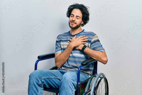 Handsome hispanic man sitting on wheelchair smiling with hands on chest with closed eyes and grateful gesture on face. health concept.