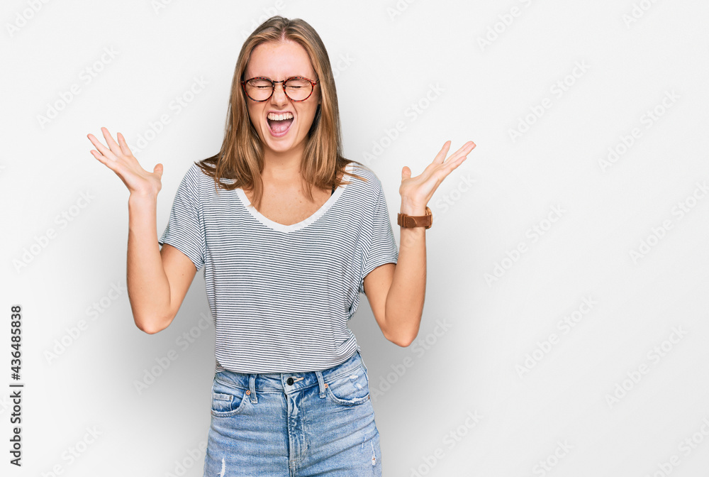 Beautiful young blonde woman wearing casual clothes and glasses celebrating mad and crazy for success with arms raised and closed eyes screaming excited. winner concept Beautiful young blonde woman wearing casual clothes and glasses celebrating mad and crazy for success with arms raised and closed eyes screaming excited. winner concept
