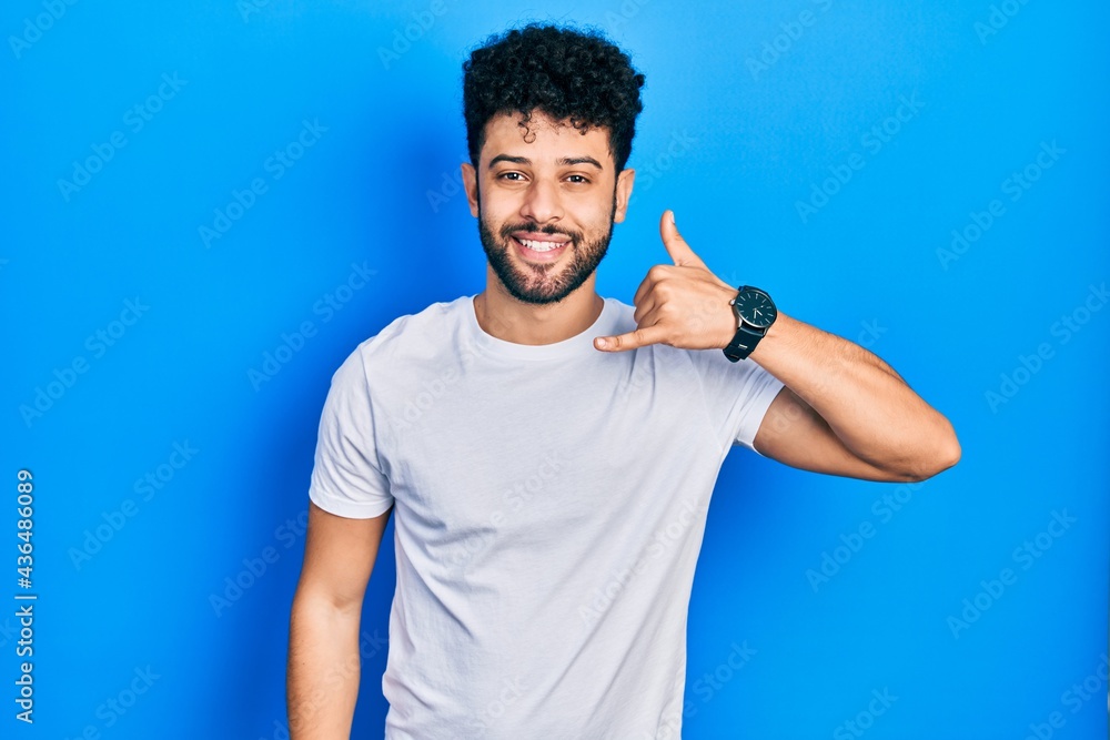 Young arab man with beard wearing casual white t shirt smiling doing phone gesture with hand and fingers like talking on the telephone. communicating concepts. Young arab man with beard wearing casual white t shirt smiling doing phone gesture with hand and fingers like talking on the telephone. communicating concepts.