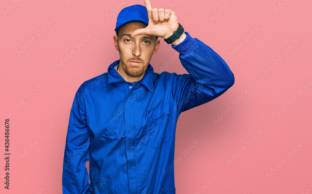 Bald man with beard wearing builder jumpsuit uniform making fun of people with fingers on forehead doing loser gesture mocking and insulting.