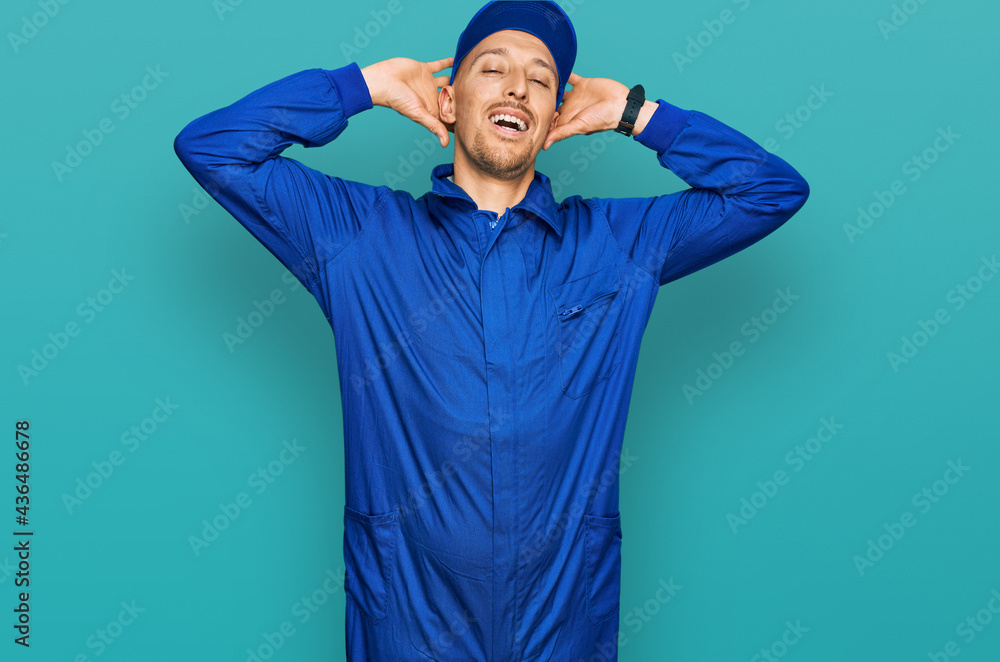 Bald man with beard wearing builder jumpsuit uniform relaxing and stretching, arms and hands behind head and neck smiling happy Bald man with beard wearing builder jumpsuit uniform relaxing and stretching, arms and hands behind head and neck smiling happy