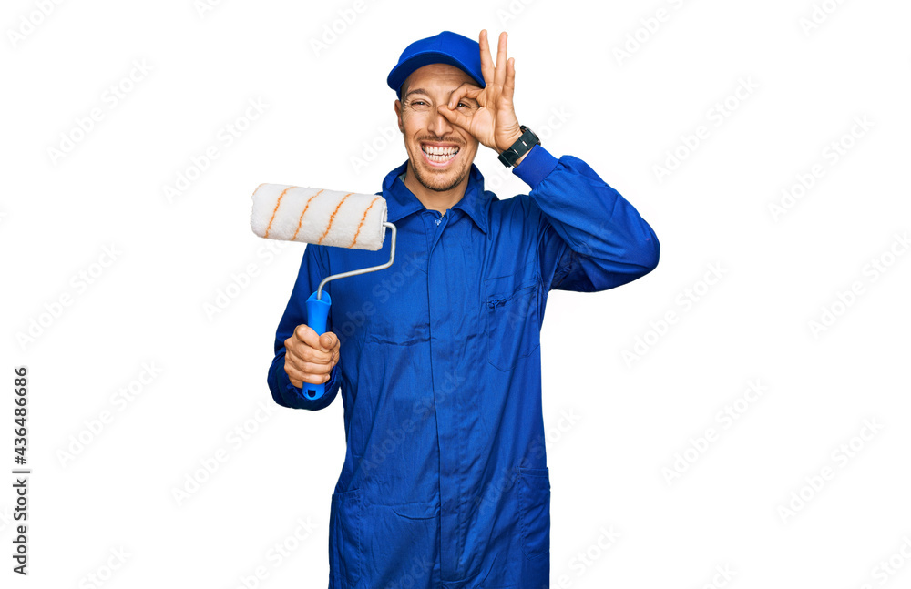 Bald man with beard holding roller painter smiling happy doing ok sign with hand on eye looking through fingers