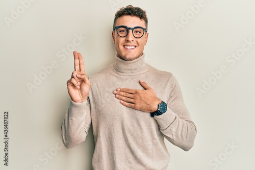 Hispanic young man wearing casual turtleneck sweater smiling swearing with hand on chest and fingers up, making a loyalty promise oath photo