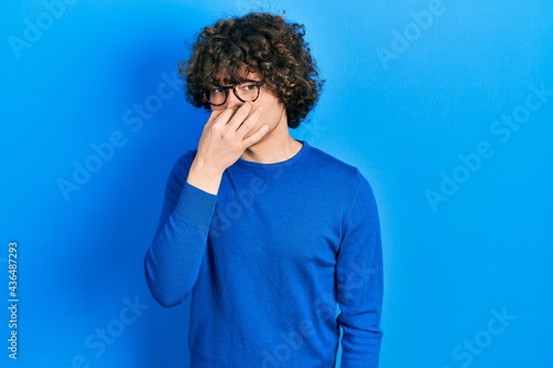 Handsome young man wearing casual clothes and glasses smelling something stinky and disgusting, intolerable smell, holding breath with fingers on nose. bad smell