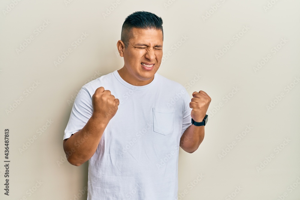 Young latin man wearing casual white t shirt excited for success with arms raised and eyes closed celebrating victory smiling. winner concept. Young latin man wearing casual white t shirt excited for success with arms raised and eyes closed celebrating victory smiling. winner concept.