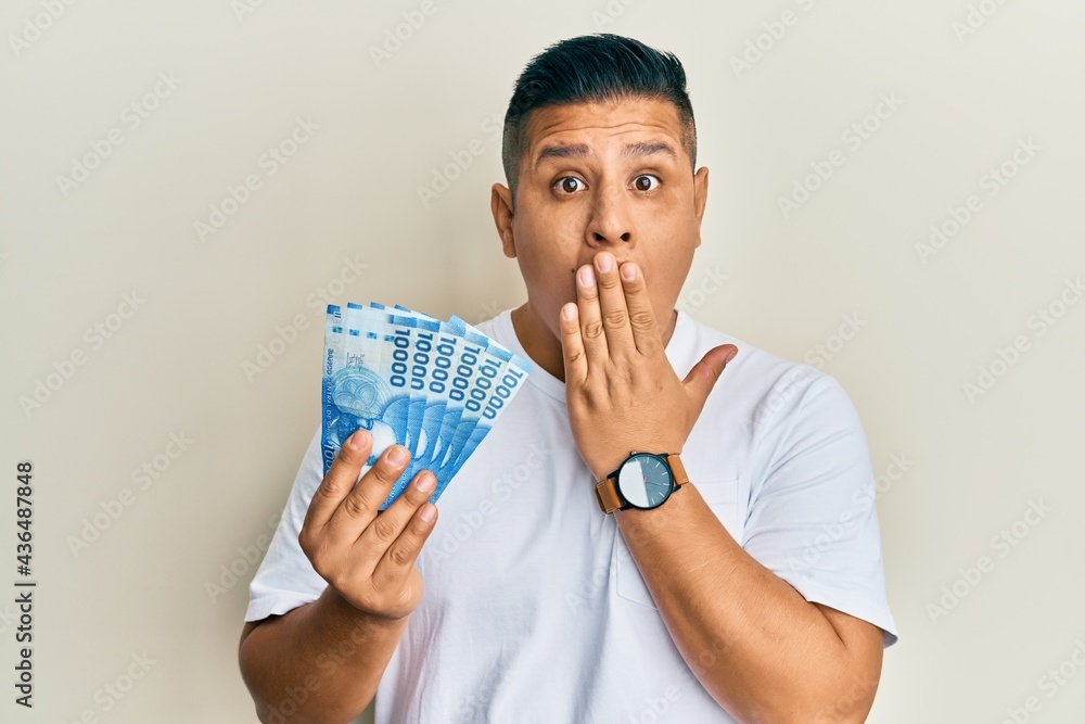 Young latin man holding 10000 chilean pesos covering mouth with hand, shocked and afraid for mistake. surprised expression Young latin man holding 10000 chilean pesos covering mouth with hand, shocked and afraid for mistake. surprised expression