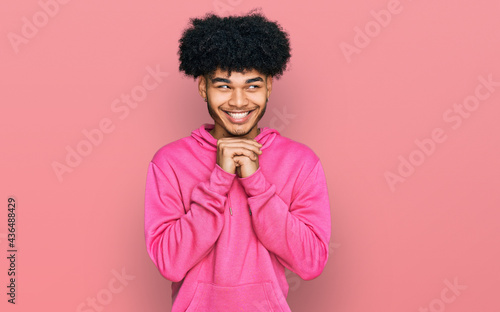 Young african american man with afro hair wearing casual pink sweatshirt laughing nervous and excited with hands on chin looking to the side