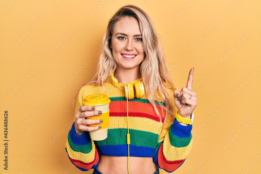 Beautiful young blonde woman drinking cup of coffee wearing headphones with a big smile on face, pointing with hand finger to the side looking at the camera. Beautiful young blonde woman drinking cup of coffee wearing headphones with a big smile on face, pointing with hand finger to the side looking at the camera.