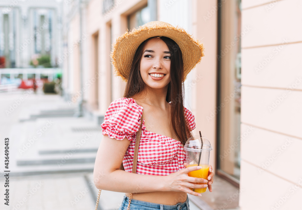 Smiling beautiful asian young woman with long hair in straw hat drinking orange juice at city street