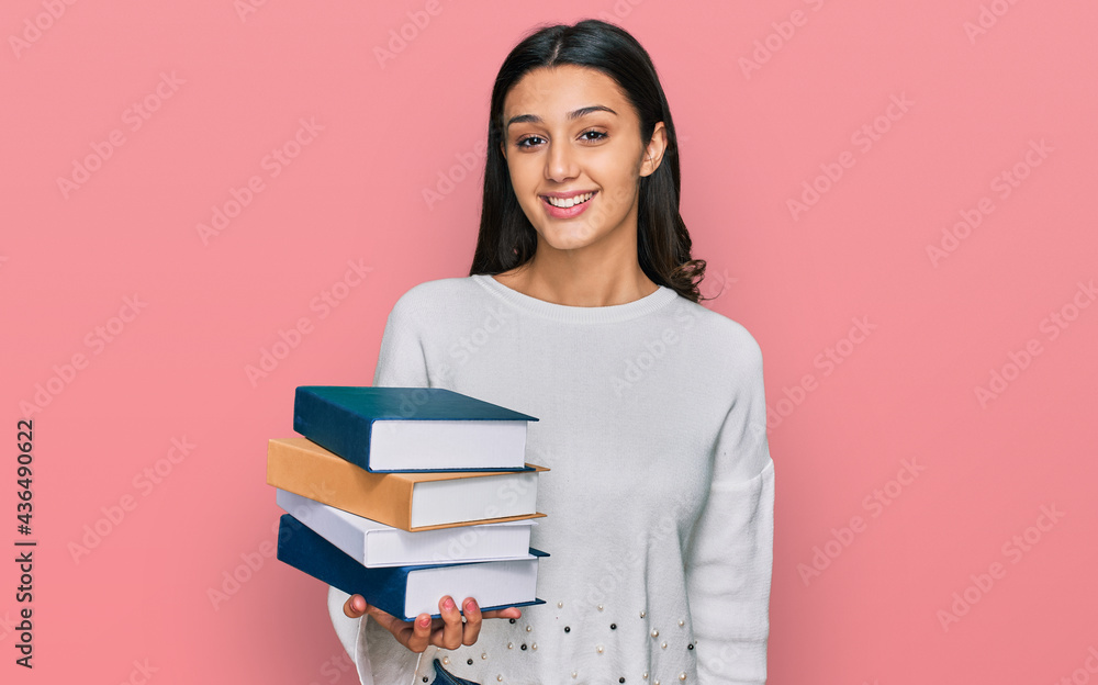 Young hispanic girl holding a pile of books looking positive and happy standing and smiling with a confident smile showing teeth Young hispanic girl holding a pile of books looking positive and happy standing and smiling with a confident smile showing teeth