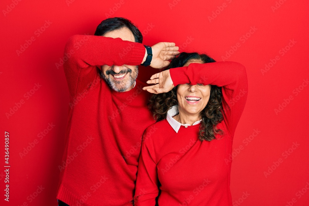 Middle age couple of hispanic woman and man hugging and standing together covering eyes with arm smiling cheerful and funny. blind concept.