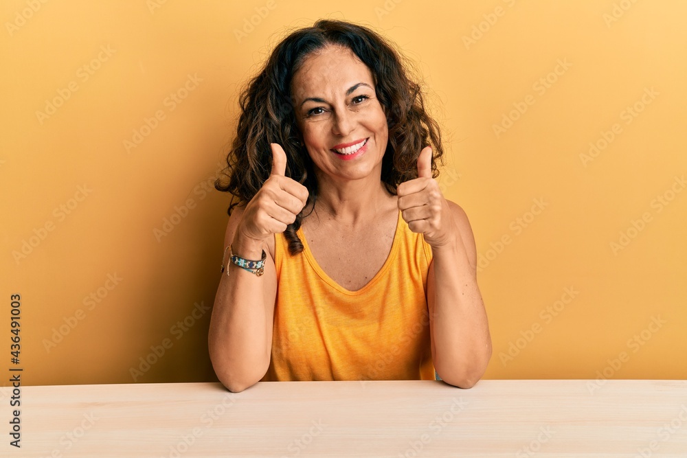 Beautiful middle age woman wearing casual clothes sitting on the table success sign doing positive gesture with hand, thumbs up smiling and happy. cheerful expression and winner gesture. Beautiful middle age woman wearing casual clothes sitting on the table success sign doing positive gesture with hand, thumbs up smiling and happy. cheerful expression and winner gesture.