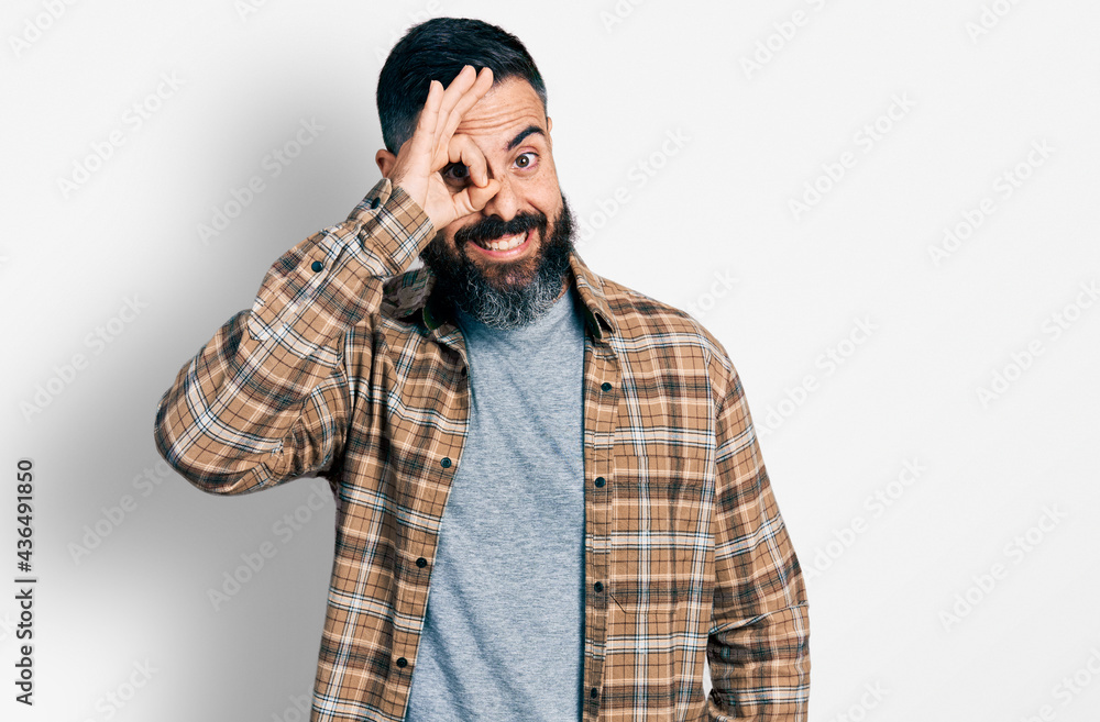 Hispanic man with beard wearing casual shirt doing ok gesture with hand smiling, eye looking through fingers with happy face. Hispanic man with beard wearing casual shirt doing ok gesture with hand smiling, eye looking through fingers with happy face.